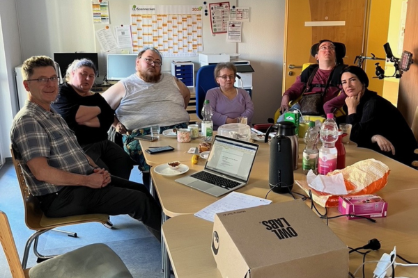 Gruppenfoto: 6 Personen sitzen an einem Tisch. Darauf ist ein Laptop, Gläser, Flaschen zu sehen. 