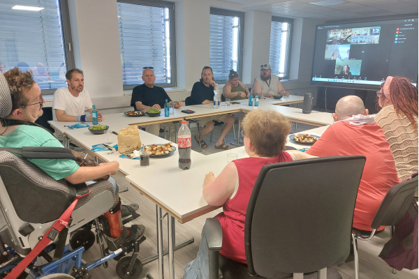 Mehrere Personen sitzen zusammen in einem Raum, auf einem Fernseher sind Zoom-Kacheln zu sehen