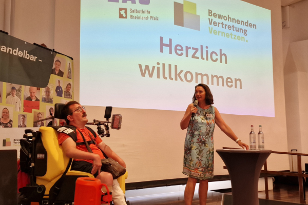 Staatssekretärin Kathrin Anklam-Trapp steht auf der Bühne und spricht in ein Mikrofon. Neben ihr Regional-Sprecher Markus Heidrich in einem E-Rollstuhl