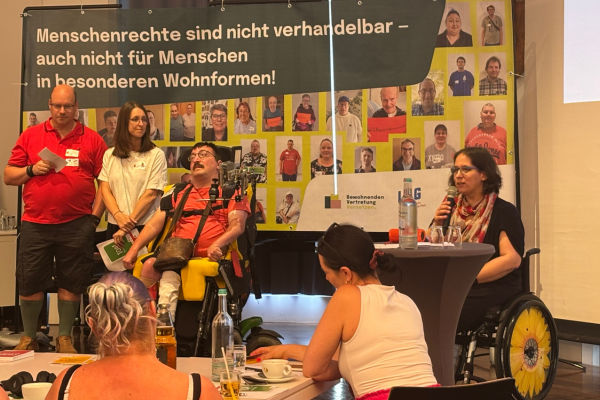 Auf der Bühne stehen Timo Gärtner, Jeanine Wein, Markus Heidrich. Die Landesbehindertenbeauftragte Ellen Kubica spricht in ein Mikrofon