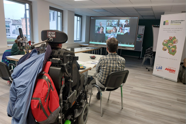 Ein hybrides Treffen: Drei Leute sitzen vor einem großen Fernseher mit weiteren digital teilnehmenden Personen