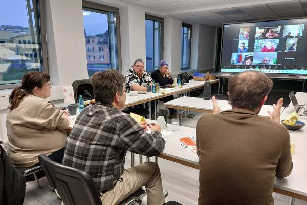 Mehrere Personen an Tischen, die auf eine Leinwand mit Zoom-Kacheln schauen