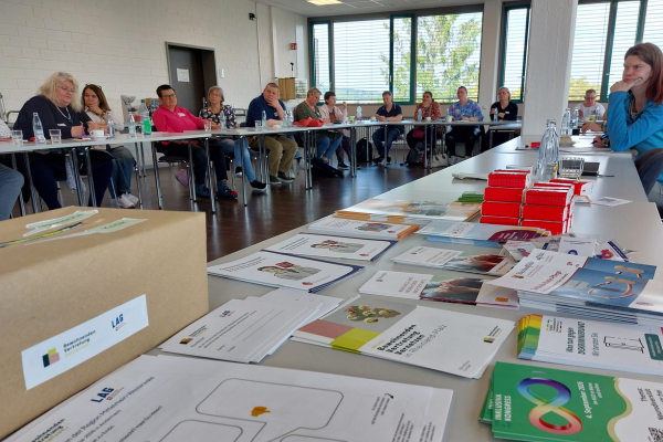 Materialien im Vordergrund - im Hintergrund Menschen die an Tischen sitzen und zuhören