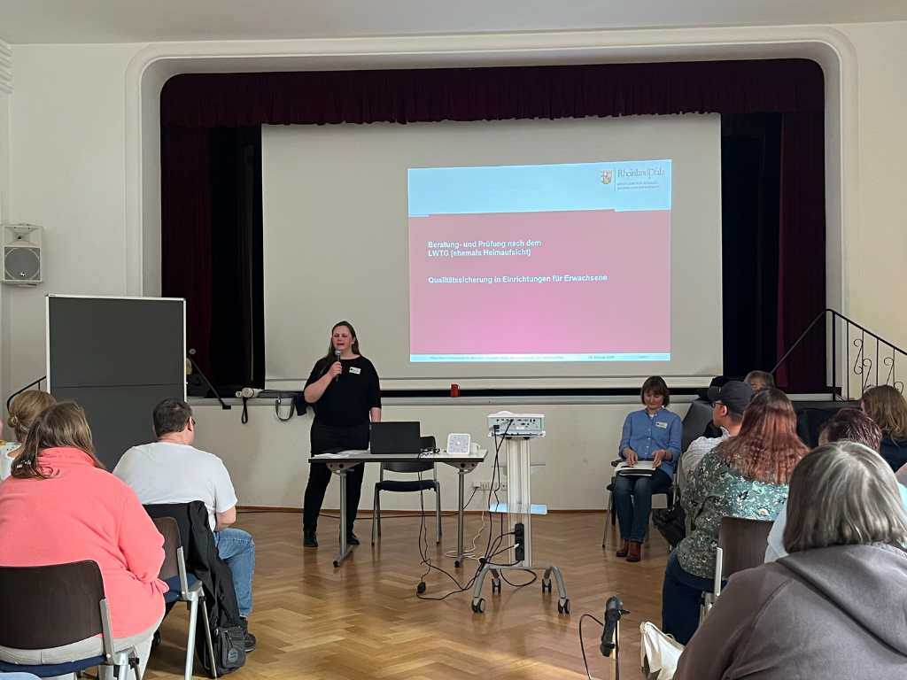 Eine Referentin steht vor einer Leinwand. Auf der Leinwand ist Text und ein Bild zu sehen. Menschen sitzen davor und hören zu.