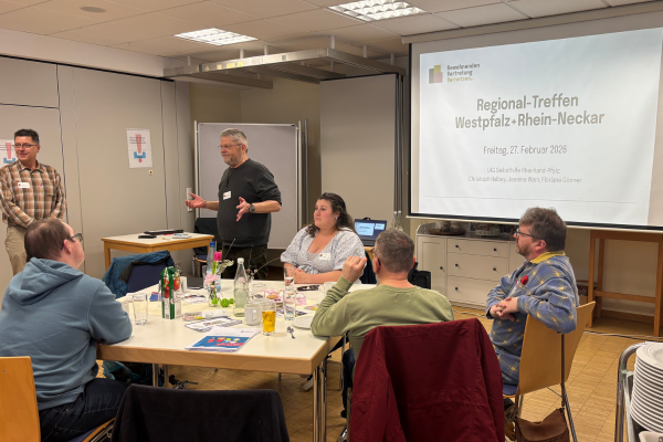 Ein Mann steht vorne vor sitzenden Menschen an Tischen. Im Hintergrund eine Beamer-Leinwand mit der Schrift: 