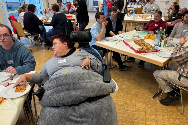 Mehrere Personen mit oder ohne sichtbare Behinderung sitzen zusammen und essen Pizza