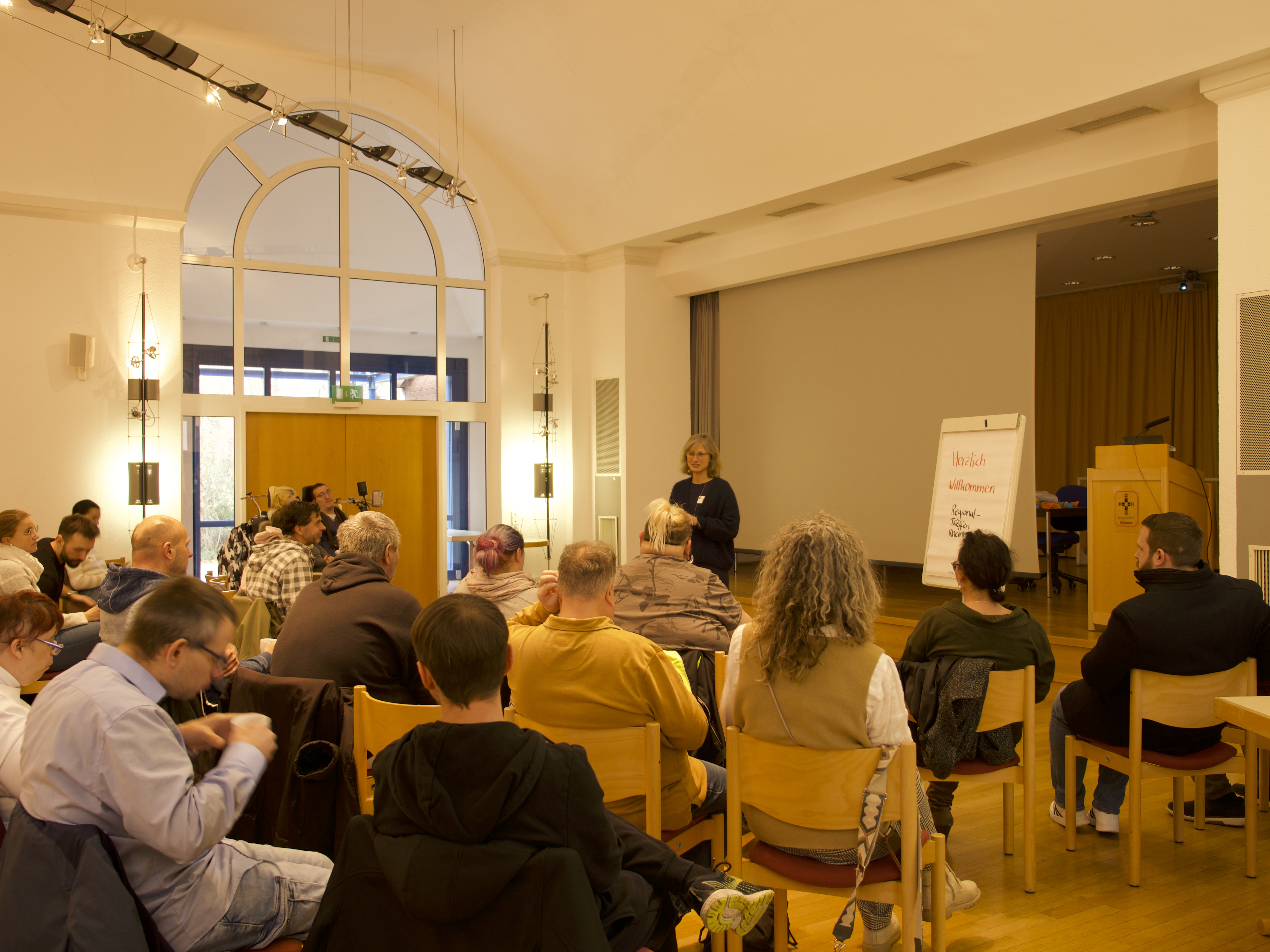 Ein hell beleuchteter Veranstaltungsraum mit hohen Decken und großen Fenstern. Eine Gruppe von Personen sitzt auf Holzstühlen und hört einer Vortragenden zu, die vor einem Flipchart steht. Auf dem Flipchart steht 'Herzlich Willkommen'. Im Hintergrund sind ein Rednerpult und eine Leinwand zu sehen.