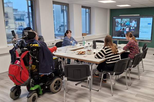 Vier Personen sitzen um einen Tisch. Im Hintergrund ist ein großer Fernseher mit den digital zugeschalteten Teilnehmenden