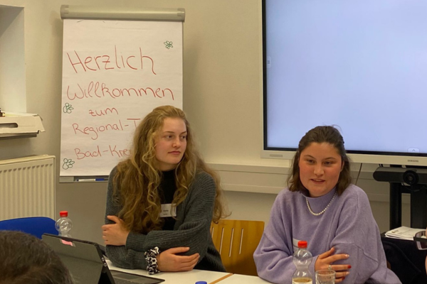 zwei junge Frauen sitzen nebeneinander vor einem Flipchart, auf dem 