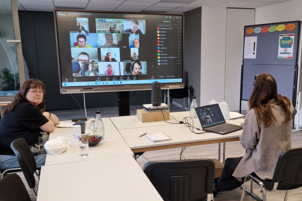 Ein hybrides Meeting: zwei Personen vor einem großen Bildschirm mit vielen Zoom-Kacheln