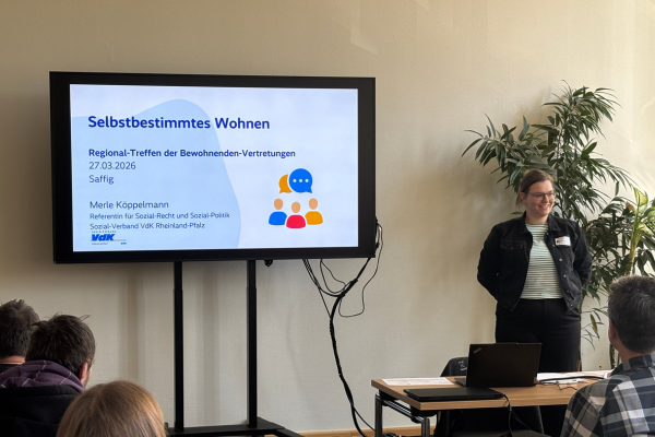 Referentin Merle Köppelmann steht neben einem Fernseher mit einer Präsentation, auf der 