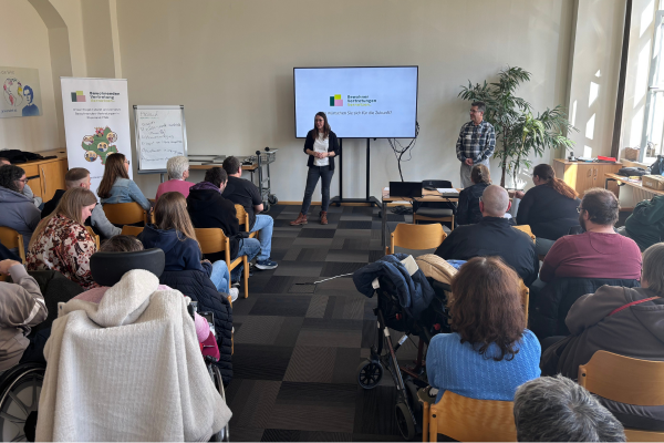 Zwei Menschen stehen neben einem Fernseher mit Präsentation. Im Vordergrund sitzen Menschen, die von hinten zu sehen sind. 