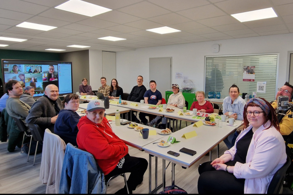 Ein hybrides Treffen: Menschen sitzen an einem Tisch, darunter Herr Speicher. Im Hintergrund sind auf einem großen Fernseher Menschen per Videokonferenz zugeschaltet