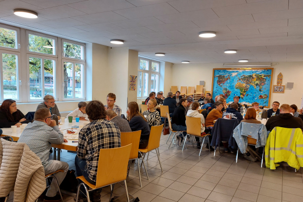 Die Aufnahme zeigt einen großen Raum mit mehreren Tischgruppen, um die herum Menschen sitzen. 